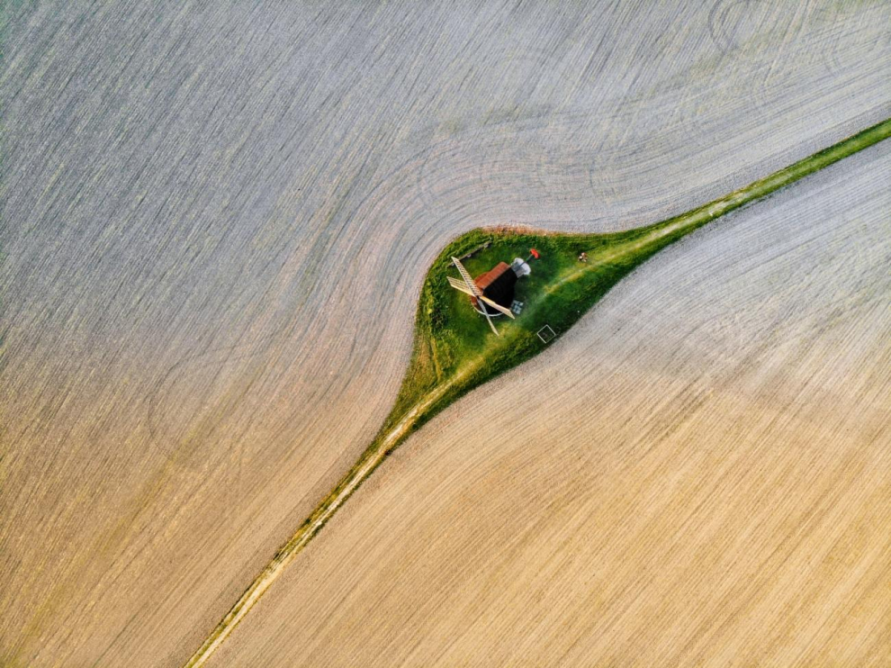 Un antiguo molino de viento situado en medio del campo en un camino de hierba. Este camino de vegetación contrasta maravillosamente con el campo circundante y desde el cielo parece una gota de agua sobre una brizna de hierba.