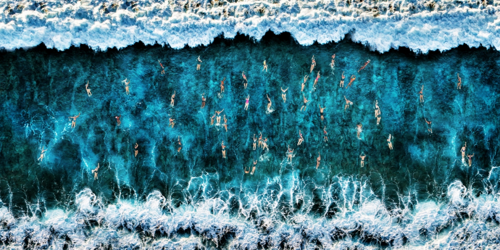 Una vista aérea de bañistas, donde el mar se convierte en el lugar de refugio, entre la alfombra azul y la espuma blanca de las olas.