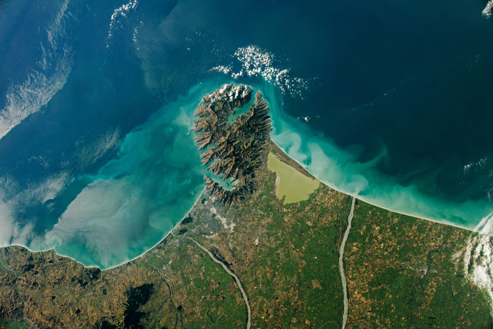 Fotografía de Christchurch y la península de Banks, una característica distintiva que sobresale de la costa este de la Isla Sur de Nueva Zelanda. Los tonos de azul a lo largo de la costa acentúan las aguas cargadas de sedimentos desde donde los ríos Waimakariri y Rakaia desembocan en el mar. La ciudad de Christchurch y el lago Ellesmere flanquean la península. Tomada el 18 de marzo de 2021