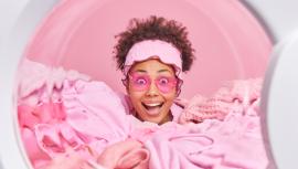 Happy surprised female housekeeper with curly hiar wears pink heart shaped sunglasses sticks head through stack of laundry. View from inside of washing machine. Housework and daily chores concept