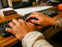 Unas manos de mujer escriben en el teclado de un ordenador, sobre una mesa de madera.