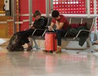 Dos personas con su equipaje esperan su vuelo sentados en un banco en el Aeropuerto de Madrid-Barajas Adolfo Suárez.
