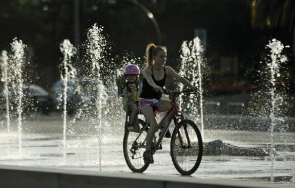 Una mujer en bici junto a una niña pasa por una fuente para refrescarse en Córdoba (Andalucía, España)